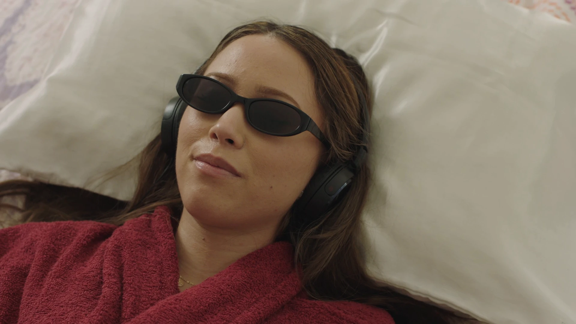 A woman in sunglasses and headphones lounges in bed in a red robe